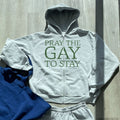 A light gray zip-up hoodie and matching sweatpants set laid flat on a wood floor, with the hoodie showing the text 'PRAY THE GAY TO STAY' in dark green. A folded blue garment is partially visible underneath on the left.